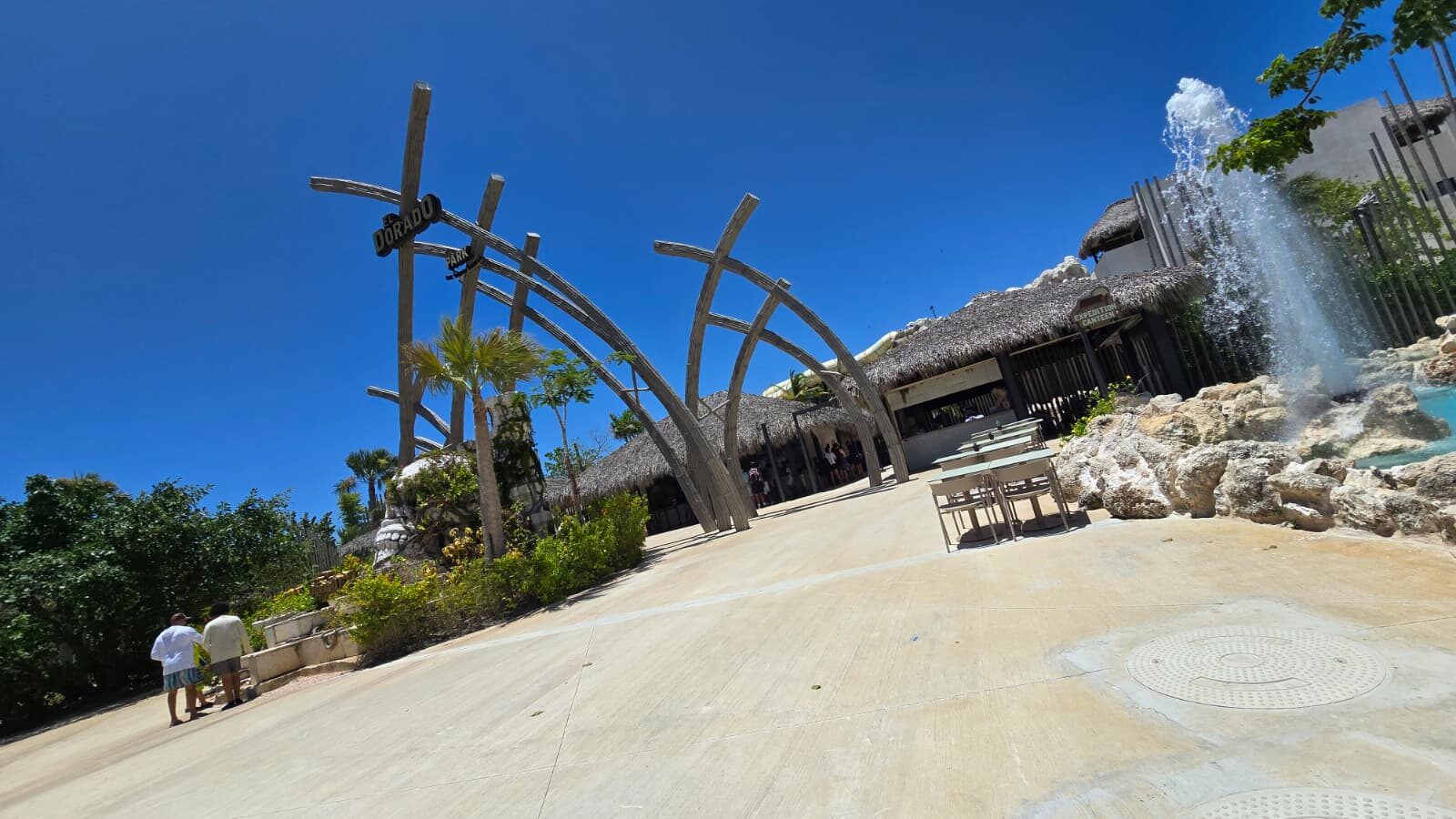 Water park entrance with wooden arches, waterfall and palm trees under a blue sky
